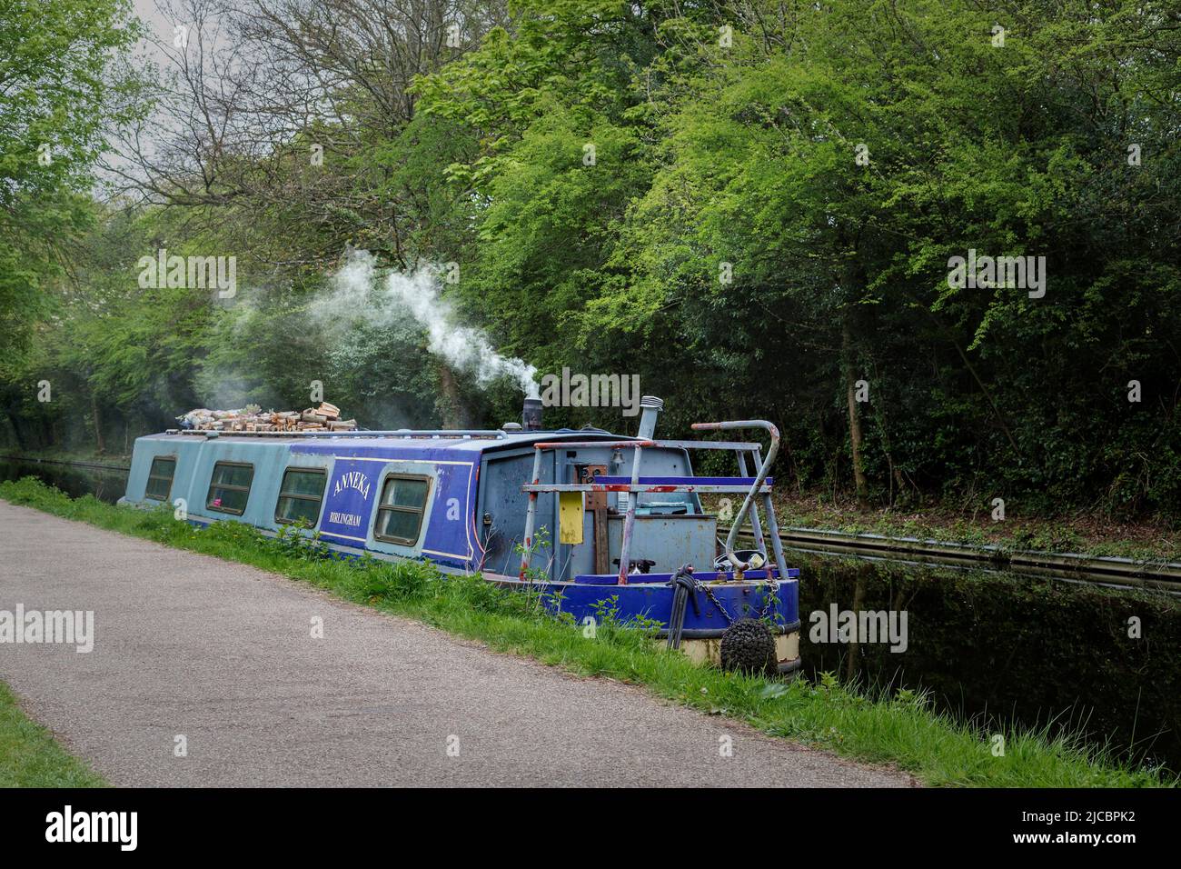 Une péniche ou un bateau étroit amarré sur un canal. Il a coupé des bûches sur le toit et la fumée provient de la cheminée d'un poêle à bois. Banque D'Images