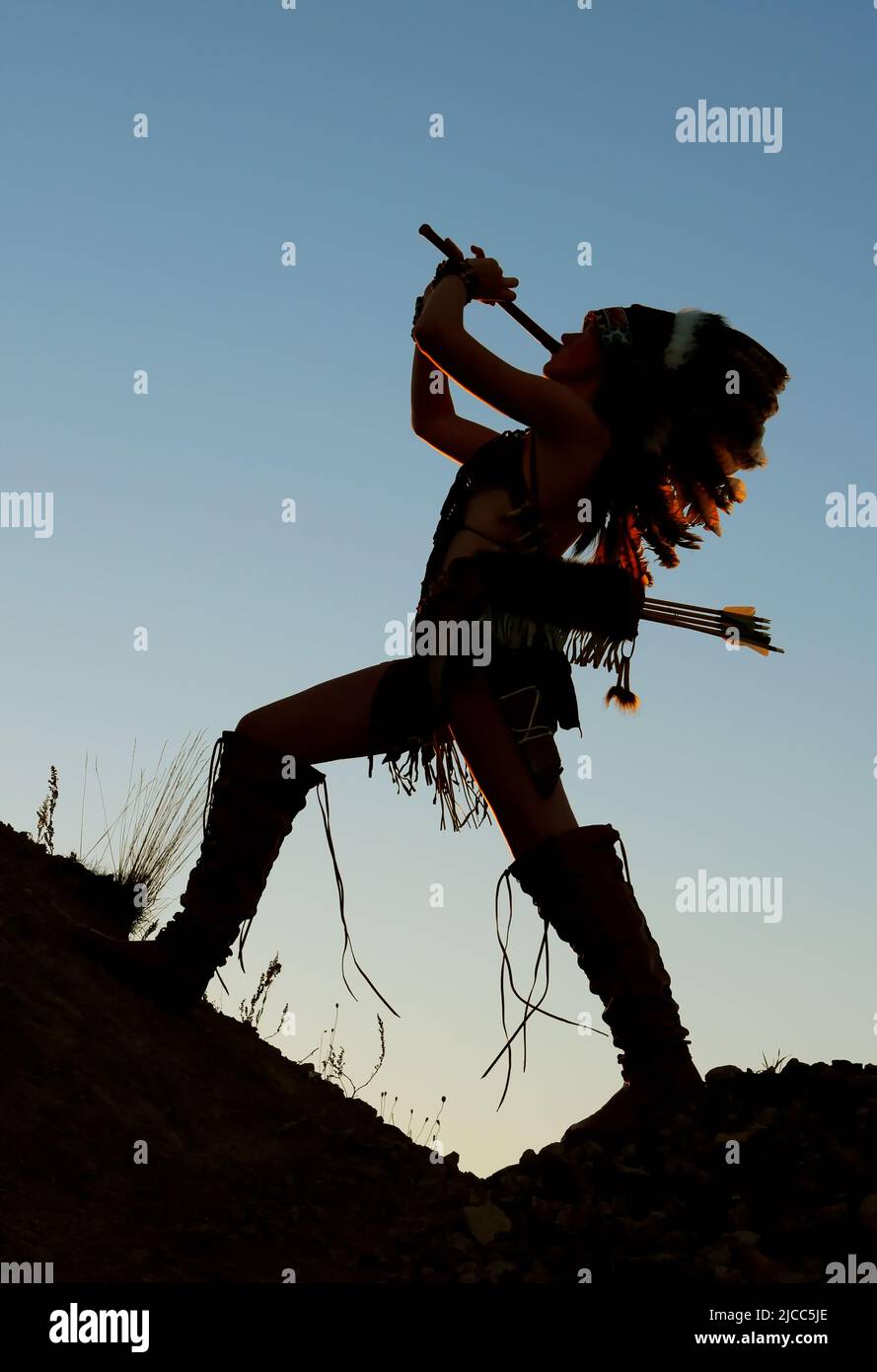 Une jeune fille indienne amérindienne joue la flûte au coucher du soleil. La fille est silhouetée comme le soleil se couche derrière elle. Banque D'Images
