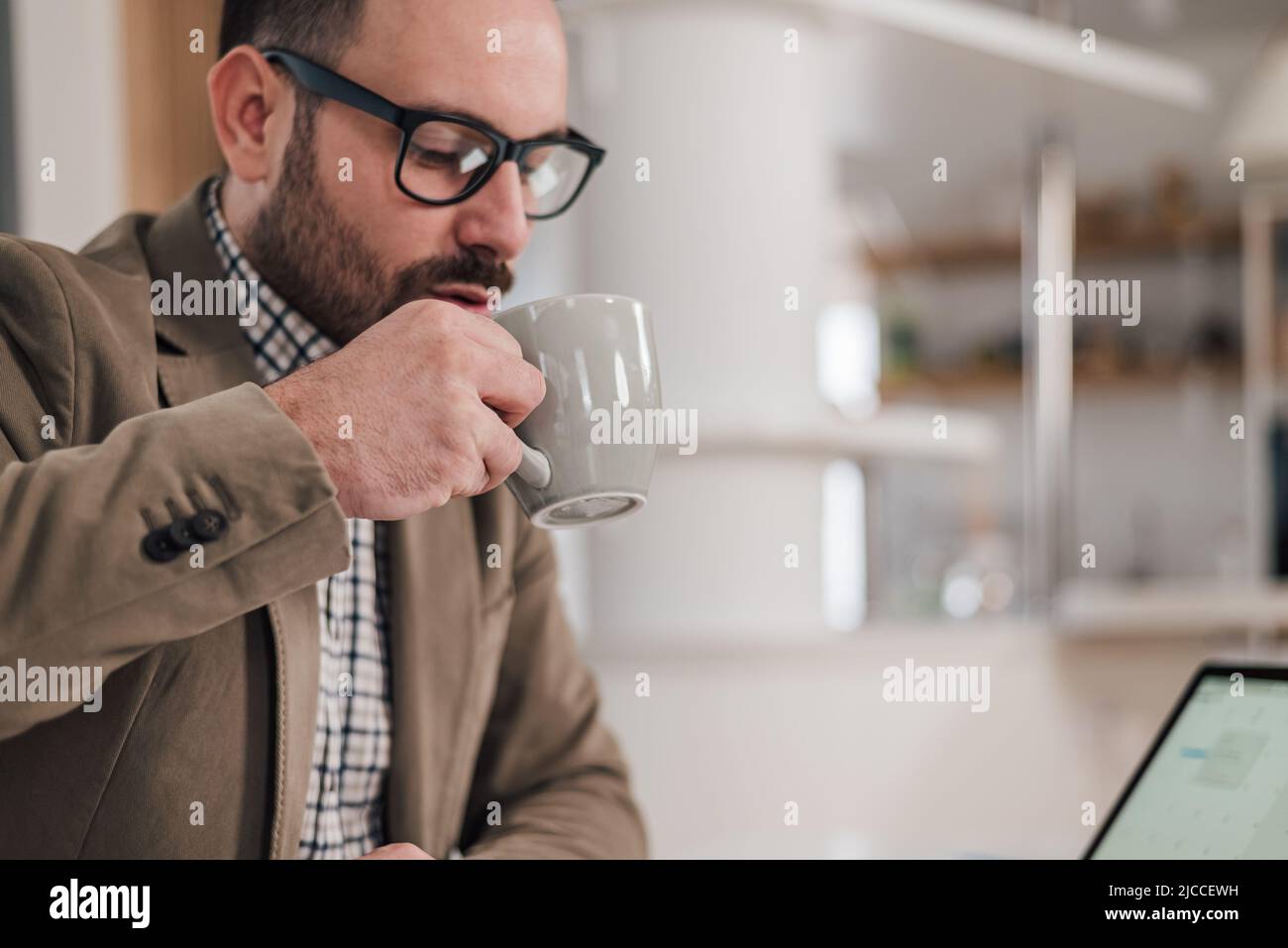 Un jeune homme d'affaires boit du café tout en travaillant. Le responsable masculin porte des lunettes. Il apprécie les rafraîchissements à son bureau. Banque D'Images