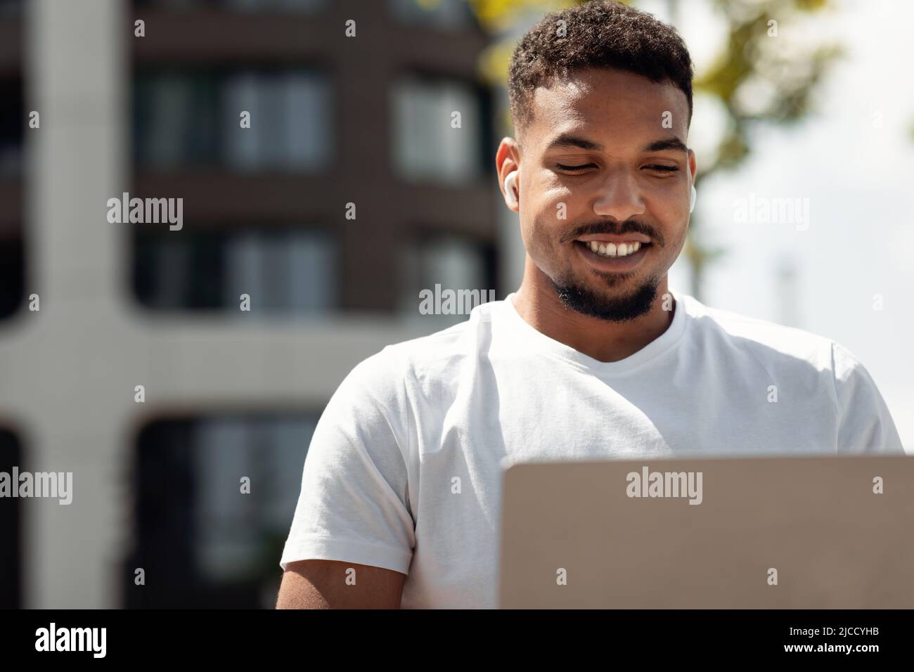 Portrait d'un homme noir freelance heureux travaillant avec un ordinateur portable à l'extérieur, assis dans la ville urbaine, espace libre Banque D'Images
