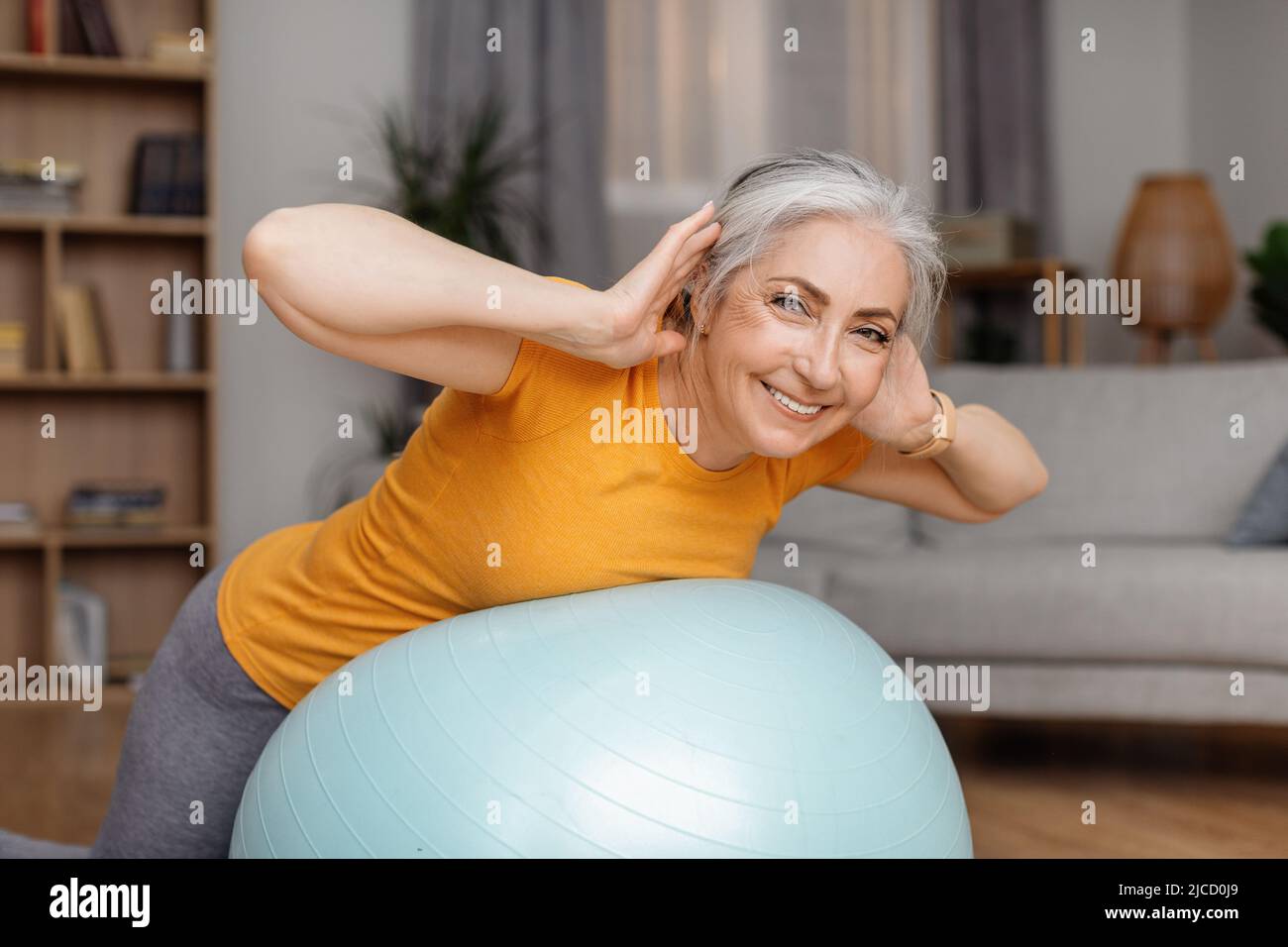 Heureuse femme senior faisant des exercices avec le ballon de fitness à la maison, la formation de ses muscles de dos pendant l'entraînement domestique Banque D'Images
