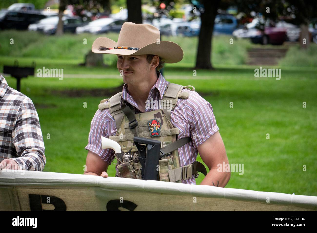 11 juin 2022, coeur d'Alene, Idaho, États-Unis : un homme armé d'un fusil et d'un revolver sourit alors qu'il tient une bannière intitulée « les groomers ne sont pas les bienvenus dans l'Idaho » lorsqu'il proteste contre la marche annuelle « Pride in the Park march » au parc McEuen, au centre-ville de coeur d'Alene, Idaho. La police de coeur d'Alene a arrêté "une petite armée" samedi de 31 membres masqués du groupe nationaliste blanc Patriot Front, qui serait "venu à l'émeute du centre-ville", selon le chef de police de coeur d'Alene. (Image de crédit : © Jacob Lee Green/ZUMA Press Wire Service) Banque D'Images