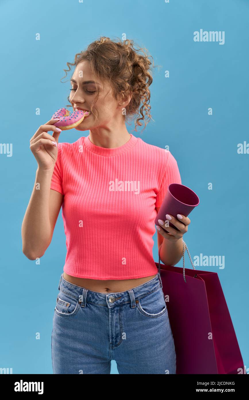 Femme maussée mangeant un beignet savoureux, tout en tenant un sac en papier dans un studio. Portrait de jolie fille appréciant le donut avec le café, ayant le repos après le shopping, isolé sur fond bleu. Concept de plaisir. Banque D'Images Femme maussée mangeant un beignet savoureux, tout en tenant un sac en papier dans un studio. Portrait de jolie fille appréciant le donut avec le café, ayant le repos après le shopping, isolé sur fond bleu. Concept de plaisir. Banque D'Images