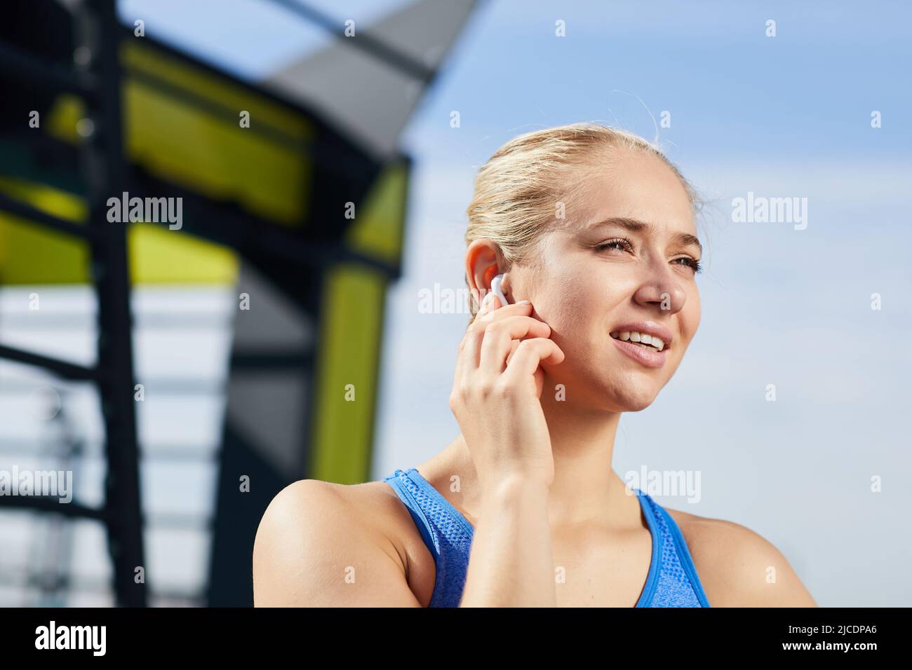 Fille sportive positive avec des cheveux blonds debout à l'extérieur et ajuster les écouteurs tout en parlant via le casque de téléphone portable Banque D'Images