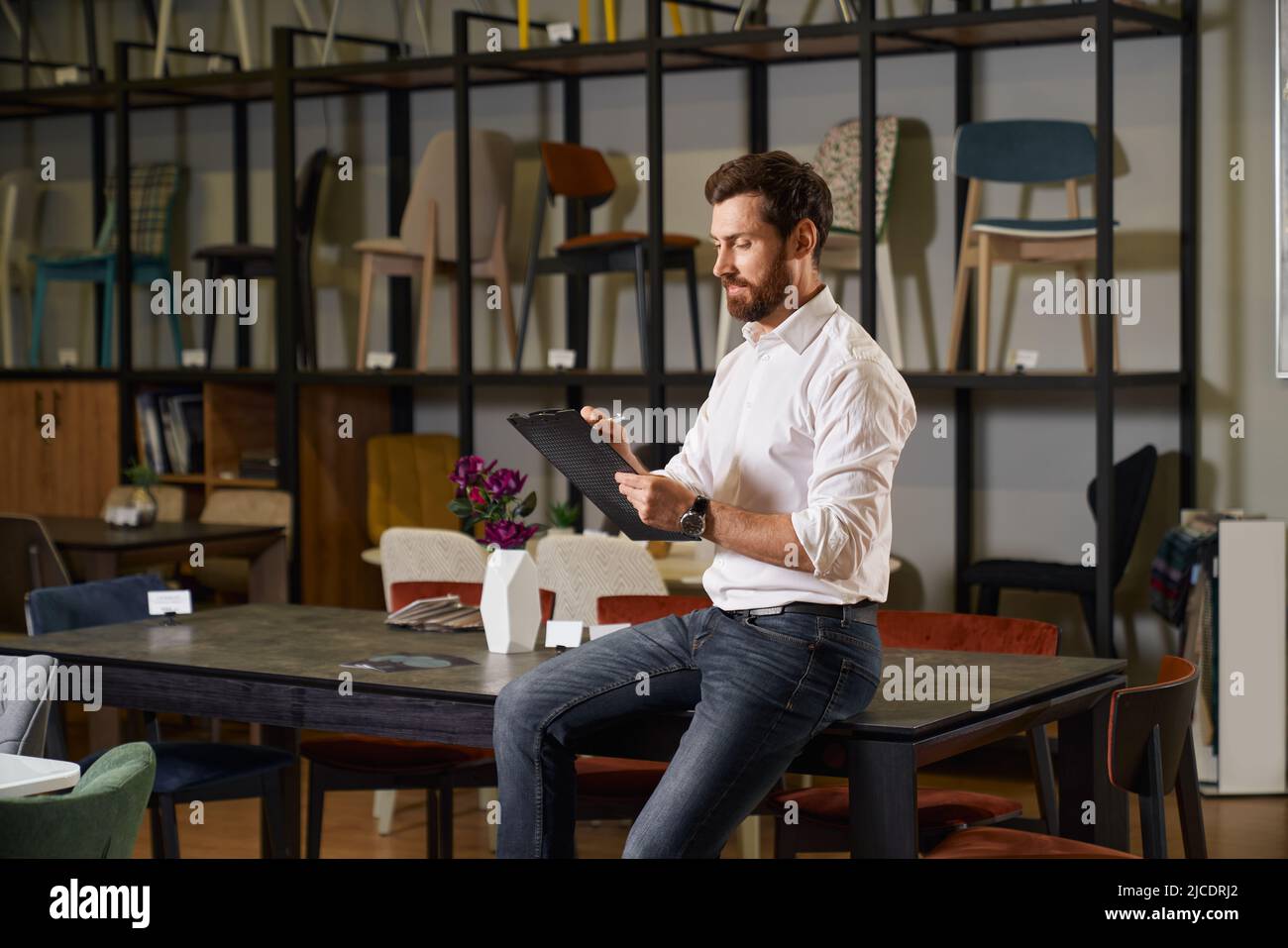 Vue de face d'un homme joyeux et concentré assis sur une table dans un magasin de meubles. Brunette homme avec la barbe écrire, regarder vers le bas, sourire, la conception. Concept de shopping et de conception. Banque D'Images Vue de face d'un homme joyeux et concentré assis sur une table dans un magasin de meubles. Brunette homme avec la barbe écrire, regarder vers le bas, sourire, la conception. Concept de shopping et de conception. Banque D'Images