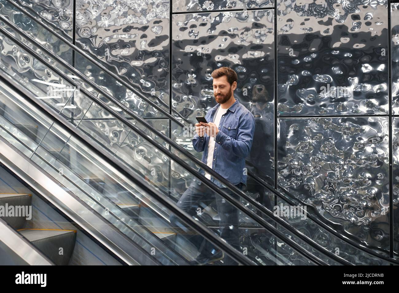 Vue avant d'un jeune homme avec une barbe debout sur un escalier roulant, tenant, en utilisant un smartphone. Homme d'affaires regardant, souriant, défilant. Concept d'affaires et de style de vie urbain moderne. Banque D'Images Vue avant d'un jeune homme avec une barbe debout sur un escalier roulant, tenant, en utilisant un smartphone. Homme d'affaires regardant, souriant, défilant. Concept d'affaires et de style de vie urbain moderne. Banque D'Images