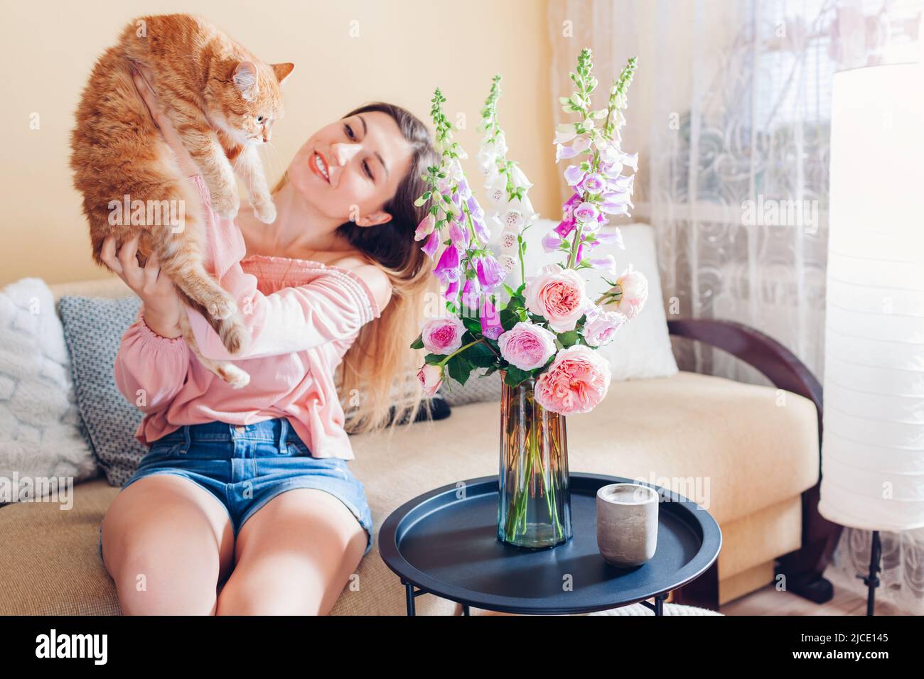 Jouer avec le chat à la maison par bouquet frais. Jeune femme assise et se détendant sur un canapé dans le salon et tenant un animal de compagnie. Confortable avec un chaton Banque D'Images