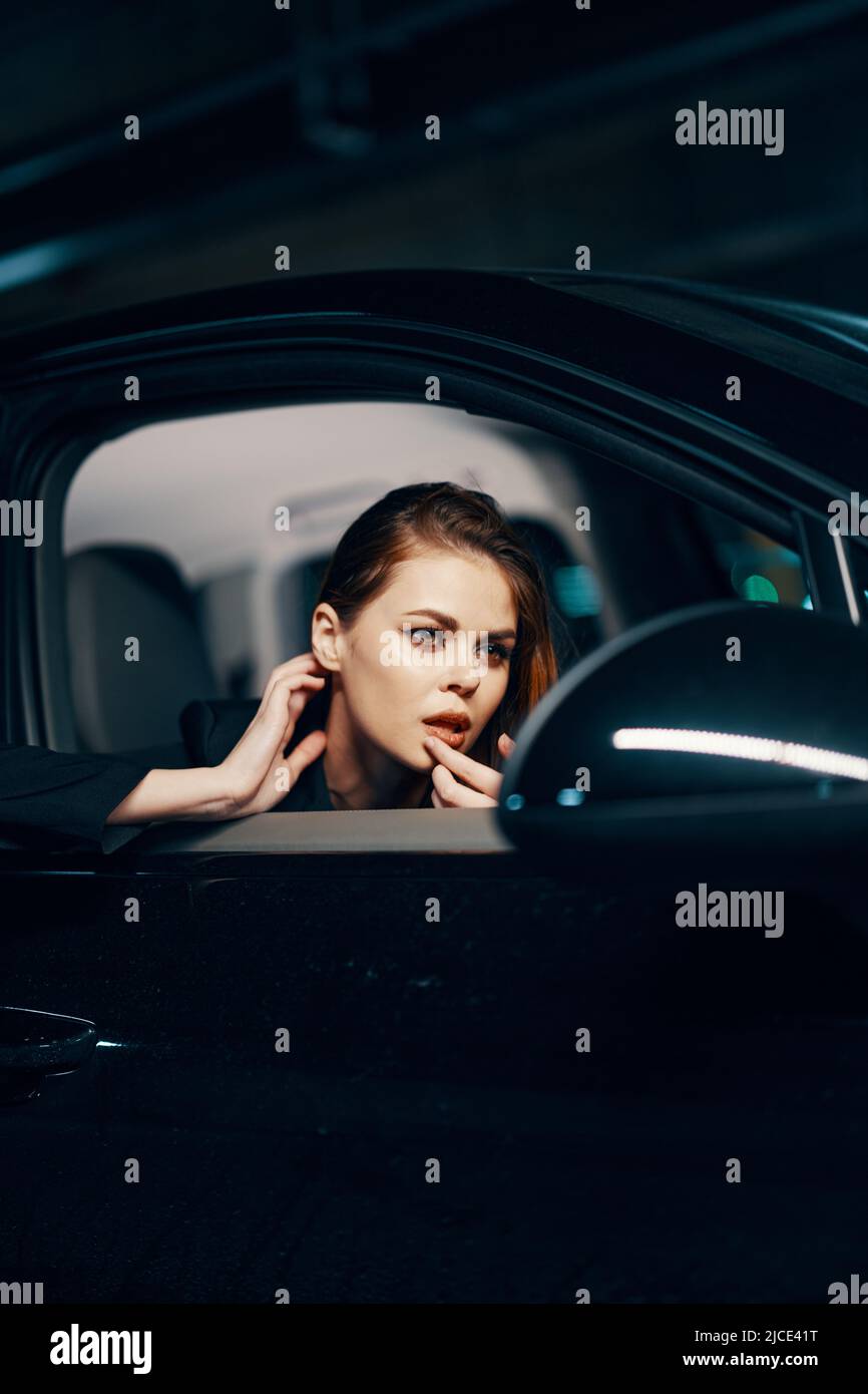 une photo verticale du côté, la nuit, d'une femme assise dans une voiture noire et regardant par la fenêtre et touchant doucement son visage tout en s'ajustant Banque D'Images