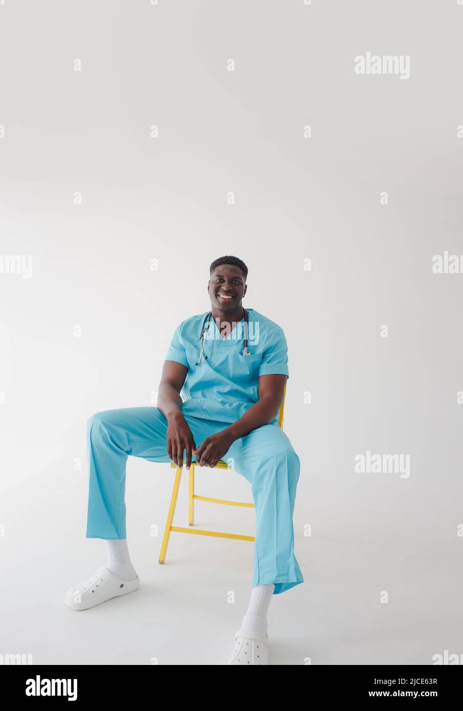 Un médecin africain dans un costume médical turquoise s'assoit sur une chaise sur un fond blanc isolé Banque D'Images