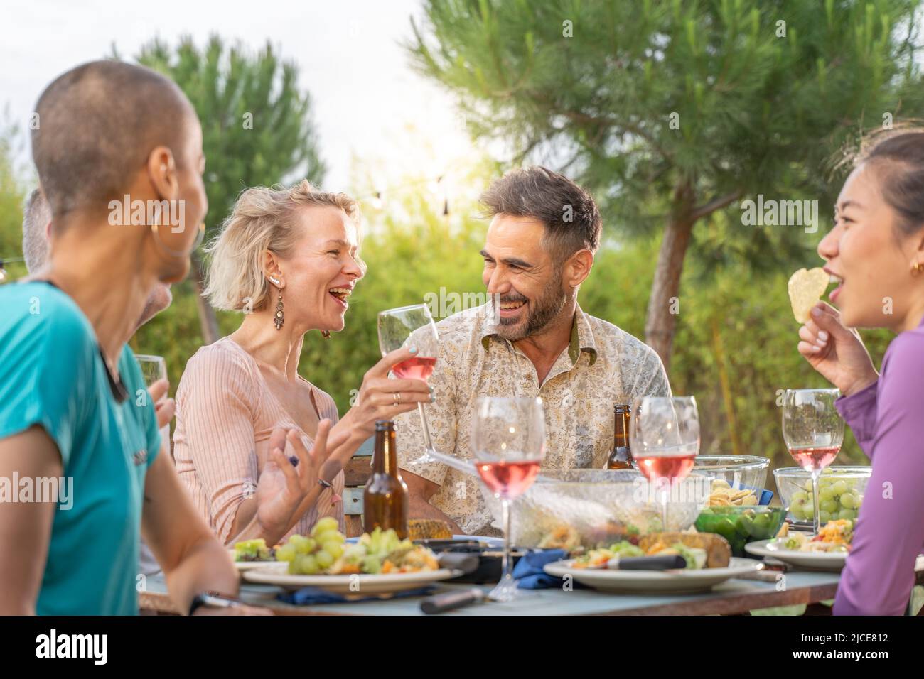 Des amis heureux rient avec un grand sourire autour de la table dans un dîner d'été. Des gens souriants mangent dans le patio de la maison. Sourires, gaieté concept. Banque D'Images