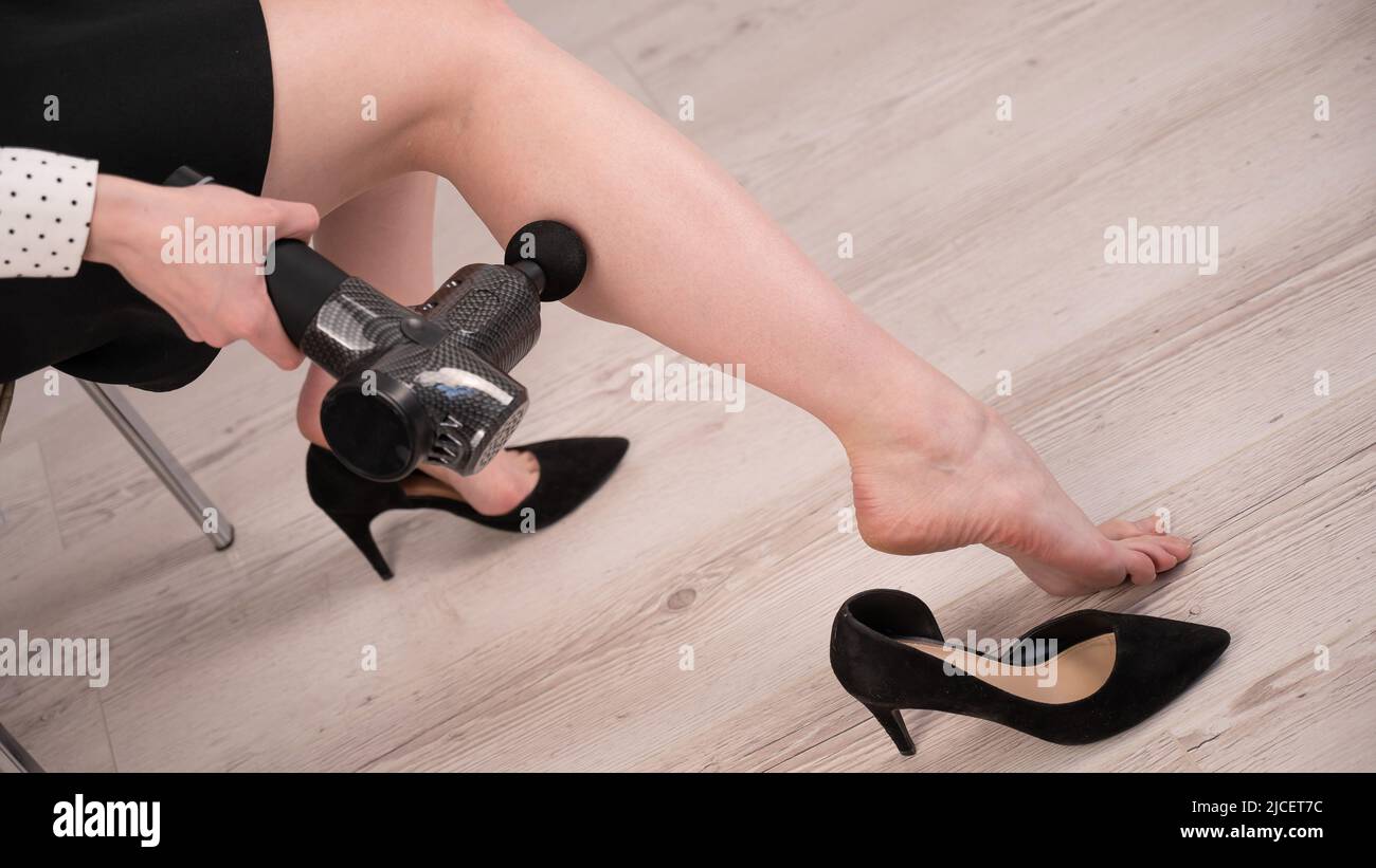 Femme d'affaires caucasienne massant sa jambe avec un masseur à percussion. Banque D'Images