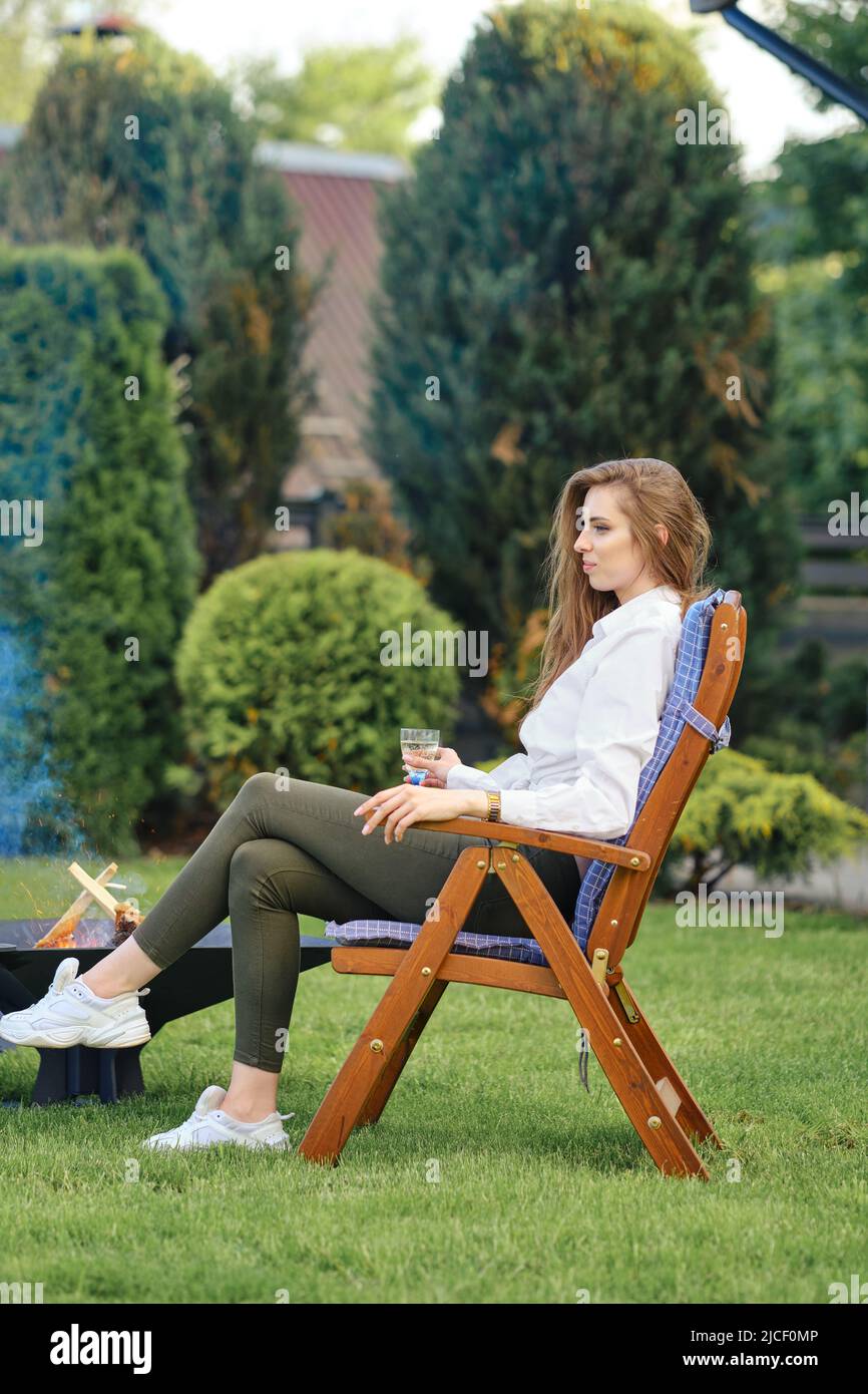 Une jeune femme assise dans une chaise longue se détend sur l'arrière-cour à côté d'un barbecue, vue dans le profil Banque D'Images