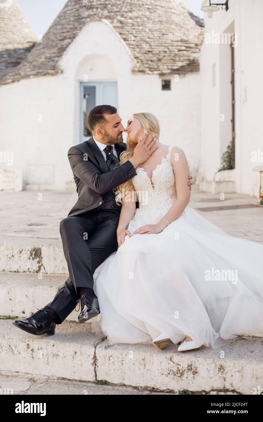 Portrait de jeune mariee blonde en robe blanche et marie brunet en costume regardant les uns les autres embrassant et assis sur les escaliers pres de la vieille maison de ville dans