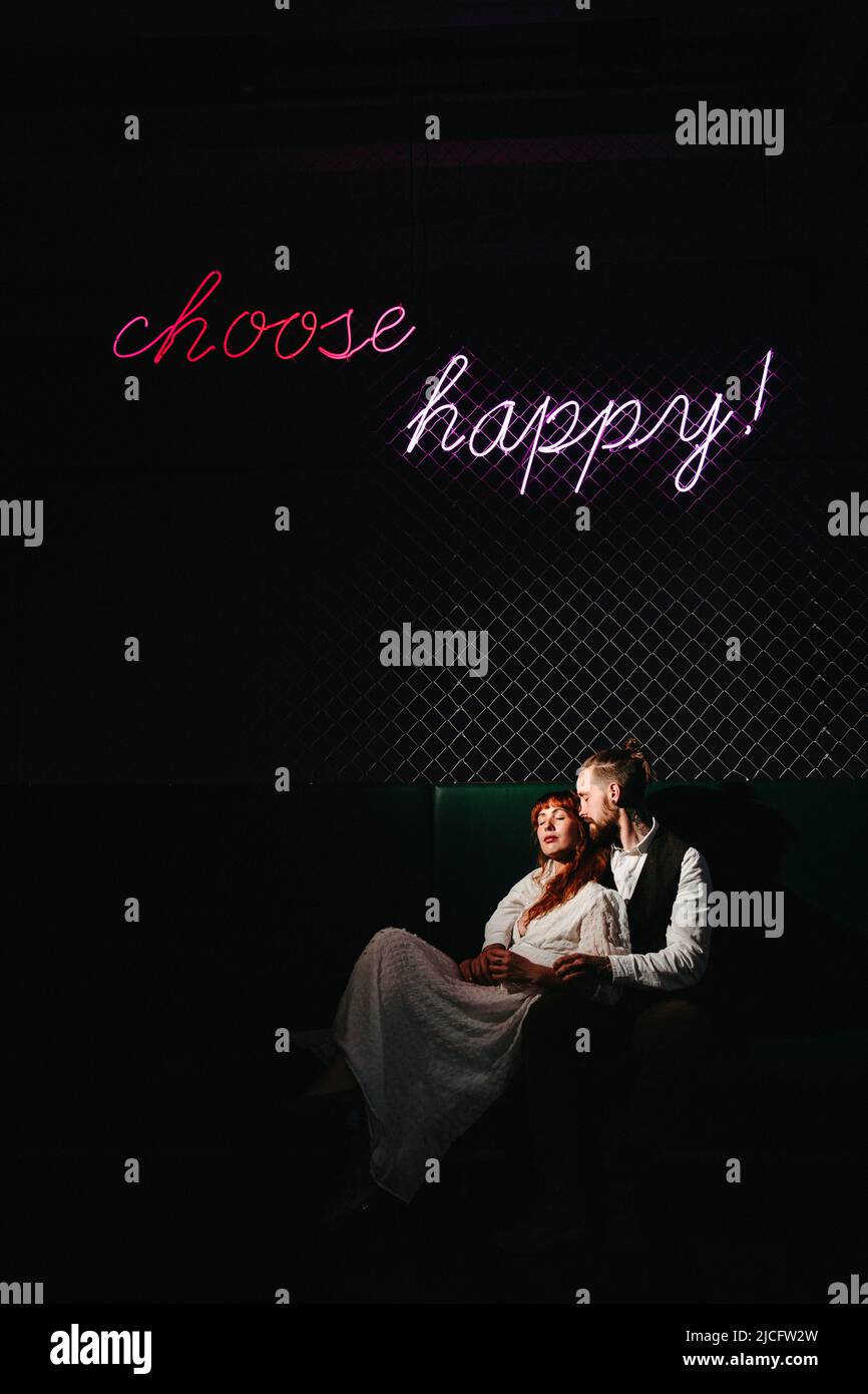 Un jeune couple amoureux assis sous un panneau au néon « Choose Happy! » Banque D'Images