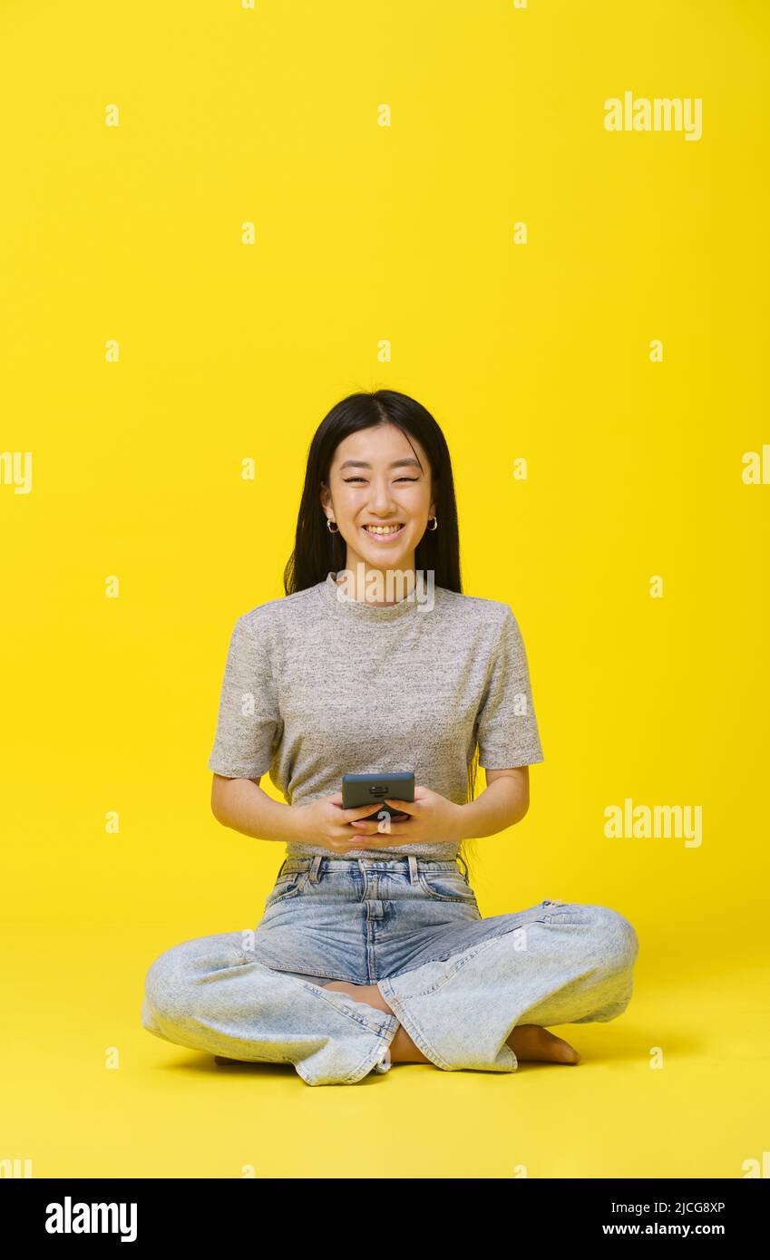 Tendre asiatique jeune fille assise sur le sol avec téléphone dans les mains texting ou shopping en ligne jeu isolé sur fond jaune. Publicité sur les applications mobiles. Copier l'espace. Banque D'Images