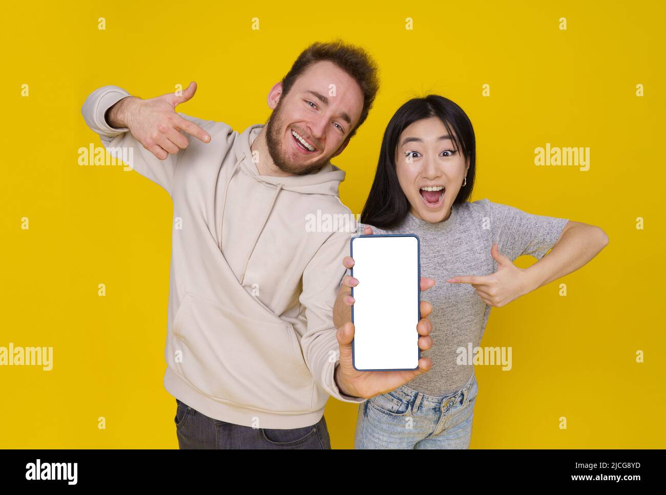 Le gars caucasien et la fille asiatique montrent les pouces vers le haut excité tenant smartphone avec blanc écran blanc, publicité d'application mobile isolée sur fond jaune. Positionnement du produit. Offre exceptionnelle. Banque D'Images