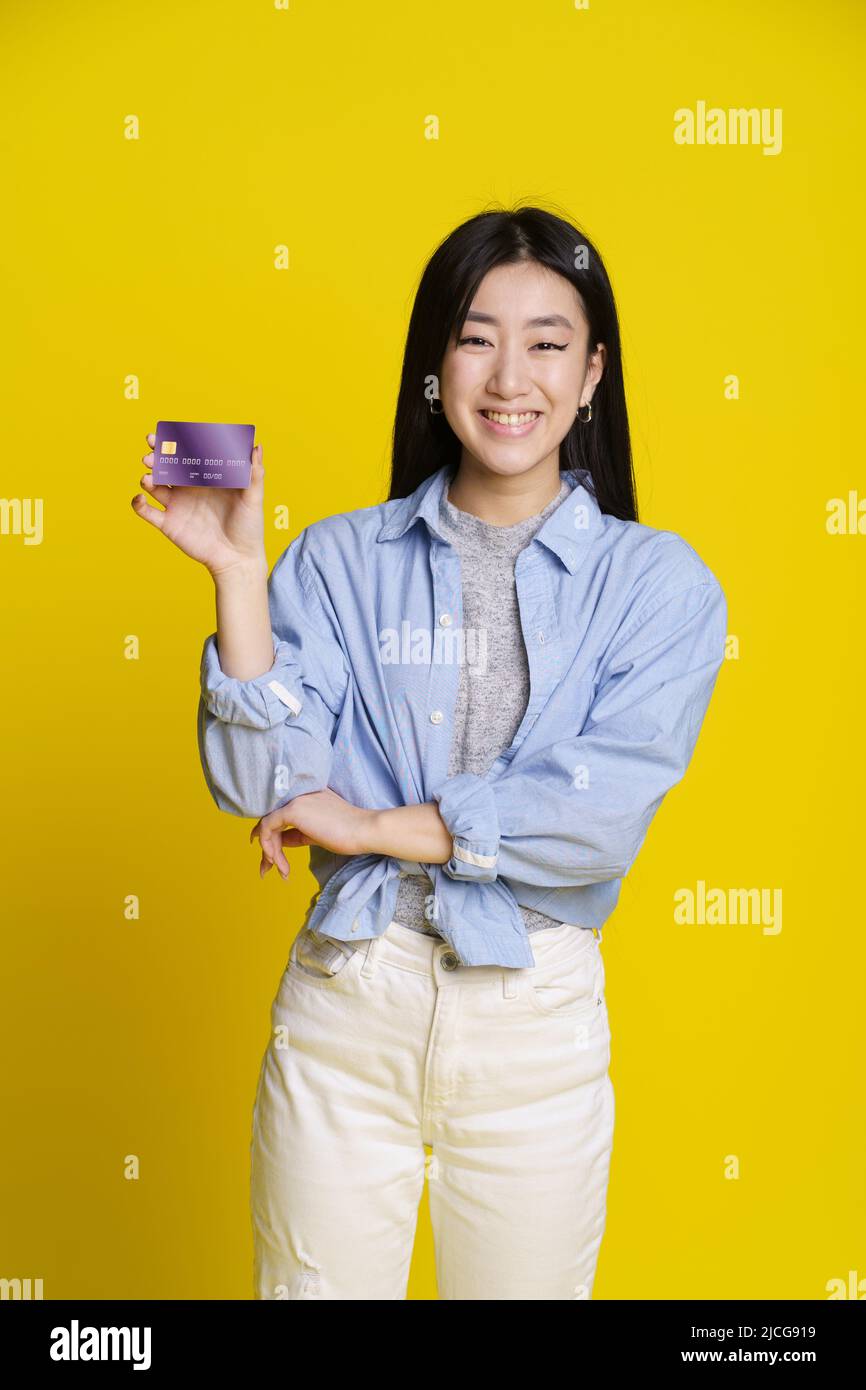 Fille asiatique souriante tenant le crédit, carte de débit à la main. Fille asiatique en chemise bleue avec carte bancaire maquette isolée sur fond jaune. Concept e-banking. Concept de commerce électronique. Banque D'Images