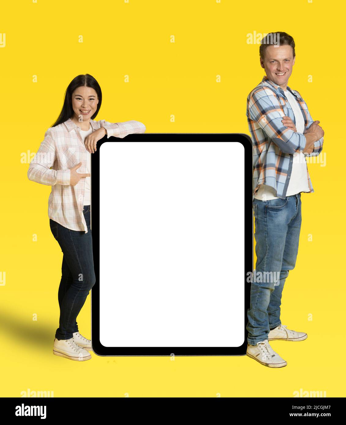 Une femme asiatique et un homme du caucase se sont pendus sur un immense Tablet pc avec écran blanc, une publicité d'application mobile regardant la caméra isolée sur fond jaune. Positionnement du produit. Banque D'Images