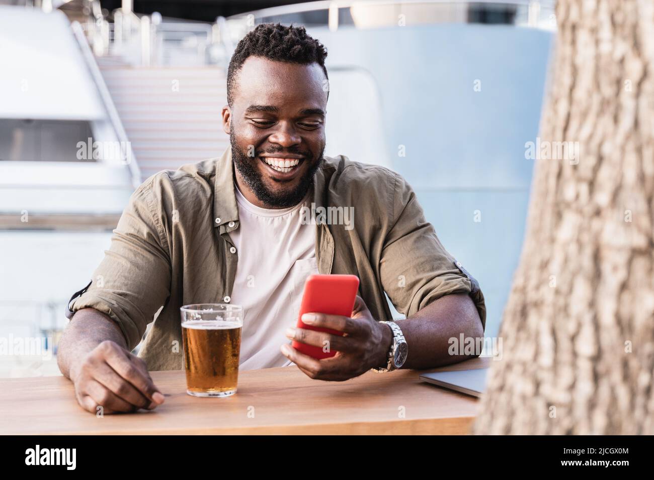 Bon homme africain qui boit de la bière au bar de la brasserie en utilisant un téléphone mobile en plein air avec un yacht de luxe en arrière-plan - Focus sur le visage Banque D'Images