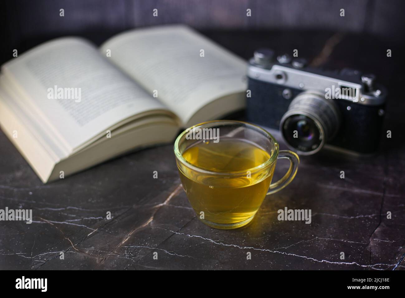 Thé vert infusé dans une tasse transparente près d'un livre ouvert et d'un appareil photo vintage sur fond sombre Banque D'Images