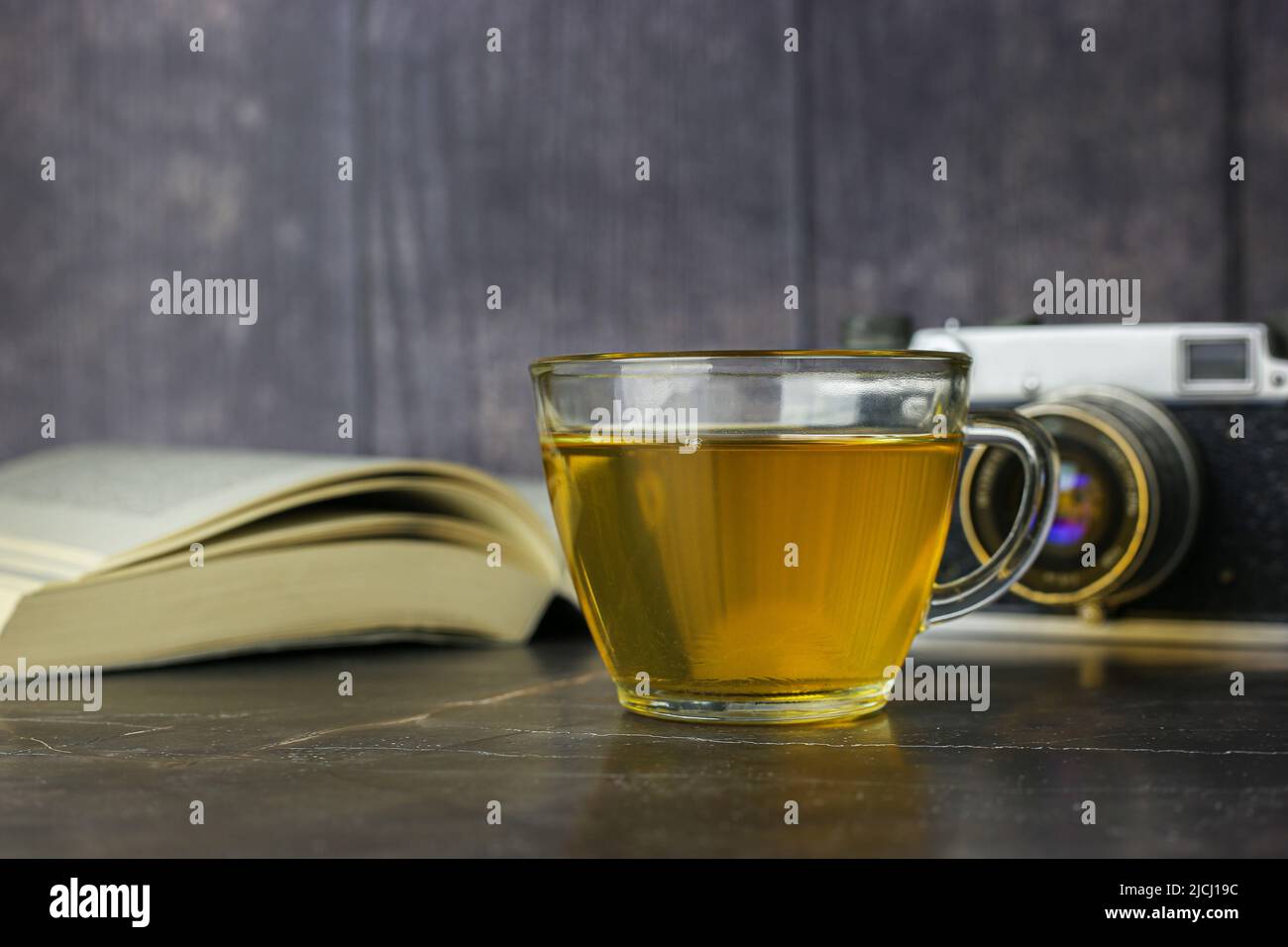 Thé vert infusé dans une tasse transparente près d'un livre ouvert et d'un appareil photo vintage sur fond sombre Banque D'Images