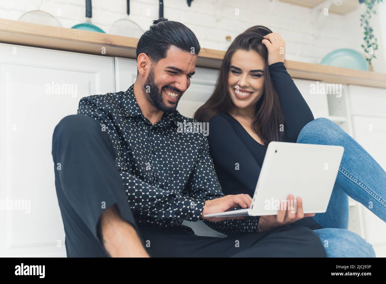 Prise en intérieur dans une cuisine moderne et lumineuse d'un jeune couple gai et interracial, tenant un ordinateur portable et riant. Photo de haute qualité Banque D'Images