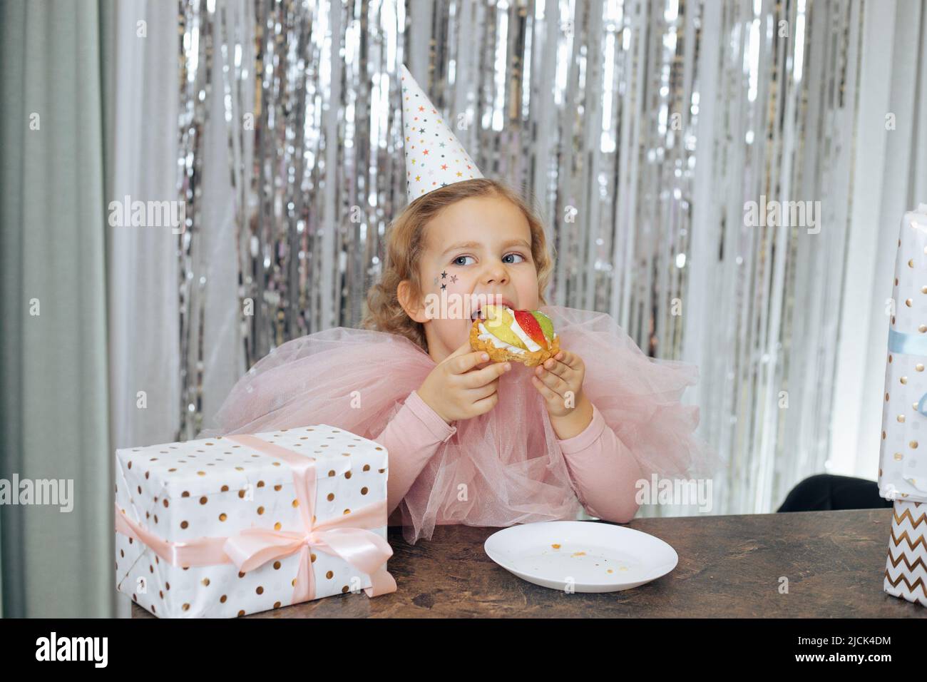 Un charmant bébé de 4 à 5 ans en chapeau de fête et une cape rose ornée de sequins décoratifs sur son visage célèbrent son anniversaire. Elle est assise attable et appréciant Banque D'Images