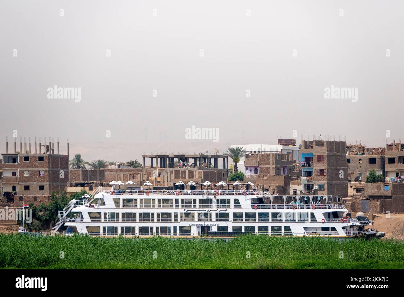 Bateau de croisière sur le Nil à bord d'un appareil photo derrière une île avec bande verte d'herbe avec mur de bâtiments derrière comme toile de fond Banque D'Images