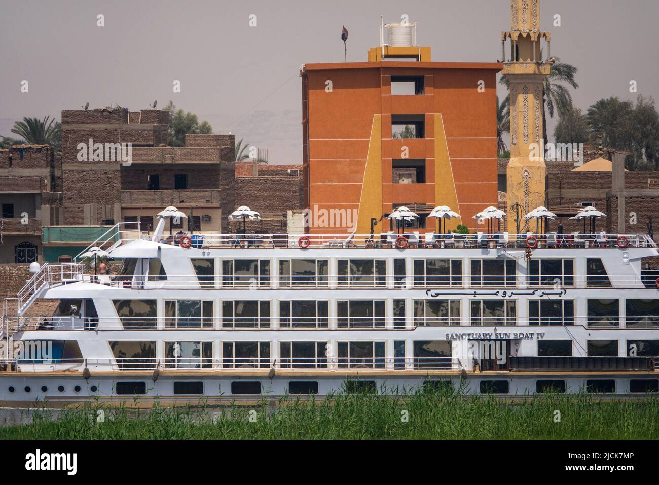 Bateau de croisière sur le Nil à bord d'un appareil photo derrière une île avec bande verte d'herbe avec mur de bâtiments derrière comme toile de fond Banque D'Images