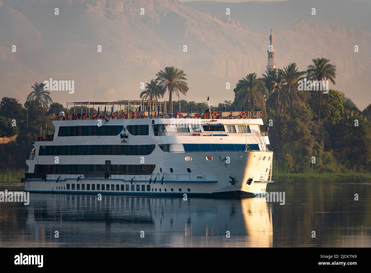 Bateau de croisière sur le Nil naviguant sur l'eau calme avec des reflets de palmiers, mosquée, montagne et navire dans la lumière du matin Banque D'Images