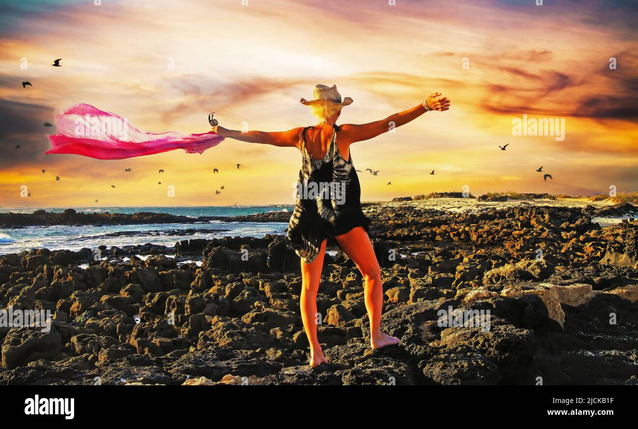 Atmosphère rocheuse mer côte plage coucher de soleil, nuages de tempête, oiseaux, une femme blonde, noir batic robe, chapeau de paille, bras étirés, écharpe rose ondulée cl Banque D'Images