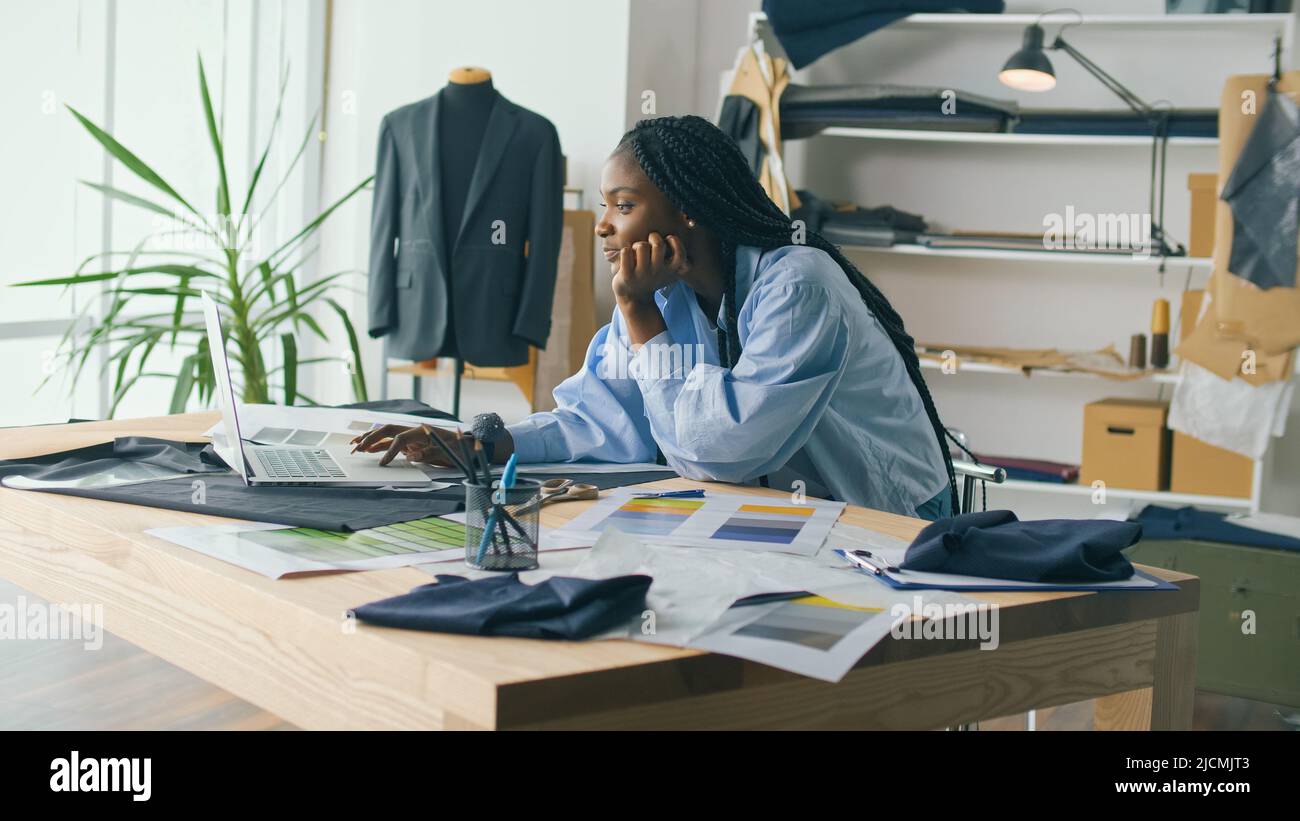 Un talentueux designer de mode afro-américain crée une nouvelle collection de mode tout en travaillant sur un ordinateur portable dans un studio Fashion Business Startup. Concept Banque D'Images