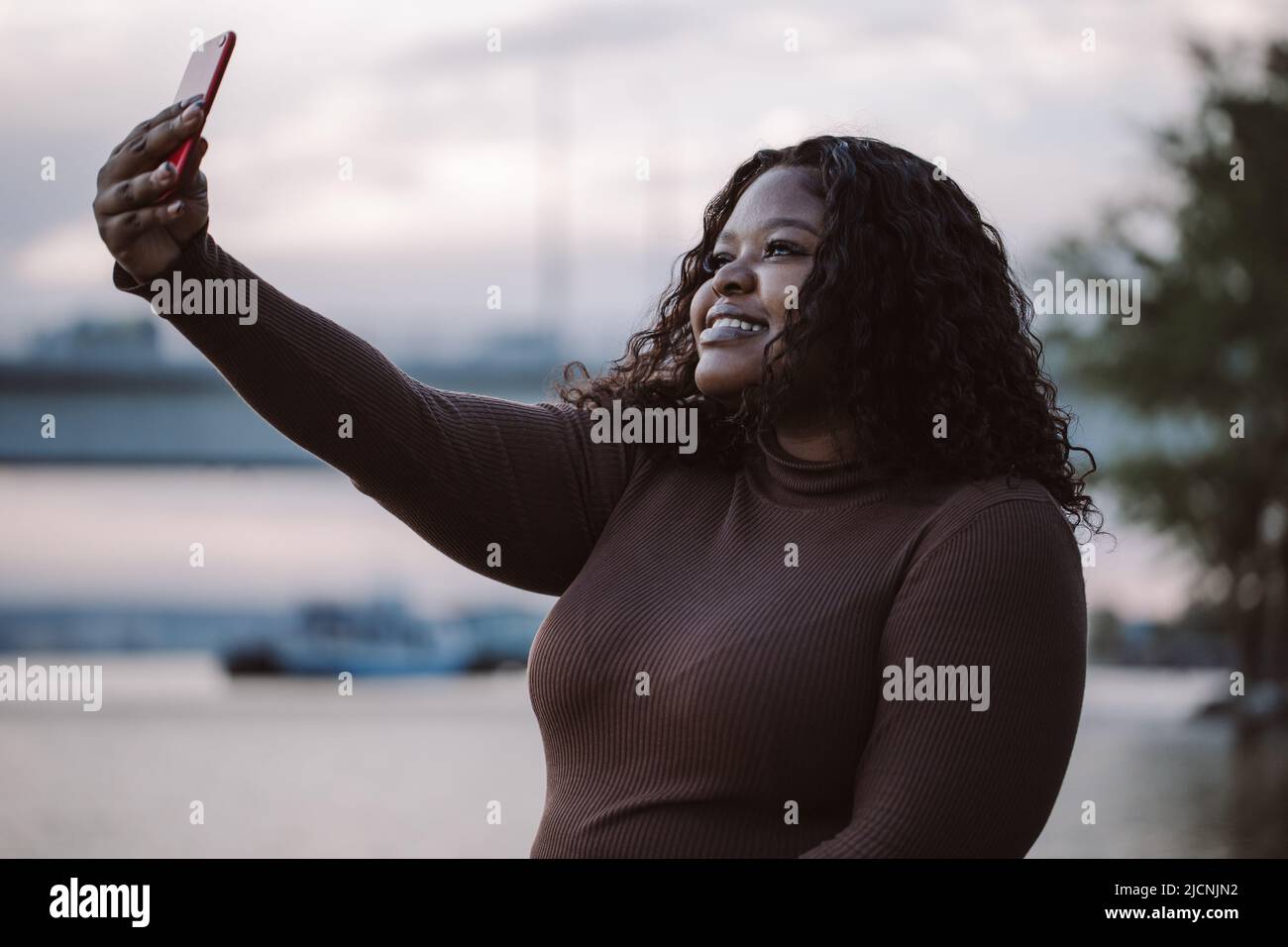 Jolie femme interraciale souriante dans une robe élégante prendre selfie, appel vidéo, photo par smartphone, se poser contre le ciel Banque D'Images