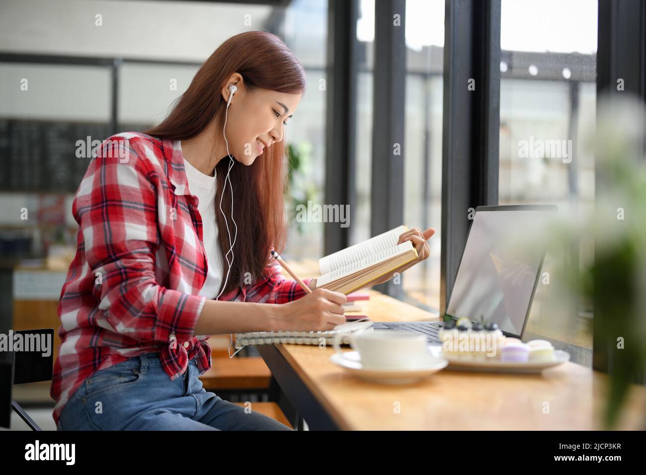 Jeune asiatique attrayante étudiante à distance travaillant dans le café, faisant ses devoirs, lisant un manuel et écoutant des cours en ligne. Banque D'Images