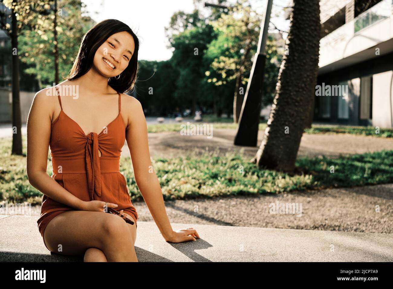 Jeune femme asiatique souriant à la caméra tout en se relaxant assis sur un banc à l'extérieur. Banque D'Images