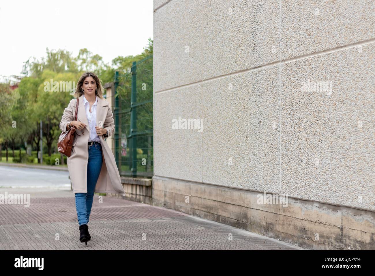 femme blonde qui va travailler dans la rue, portant un manteau beige, une chemise blanche et un jean Banque D'Images