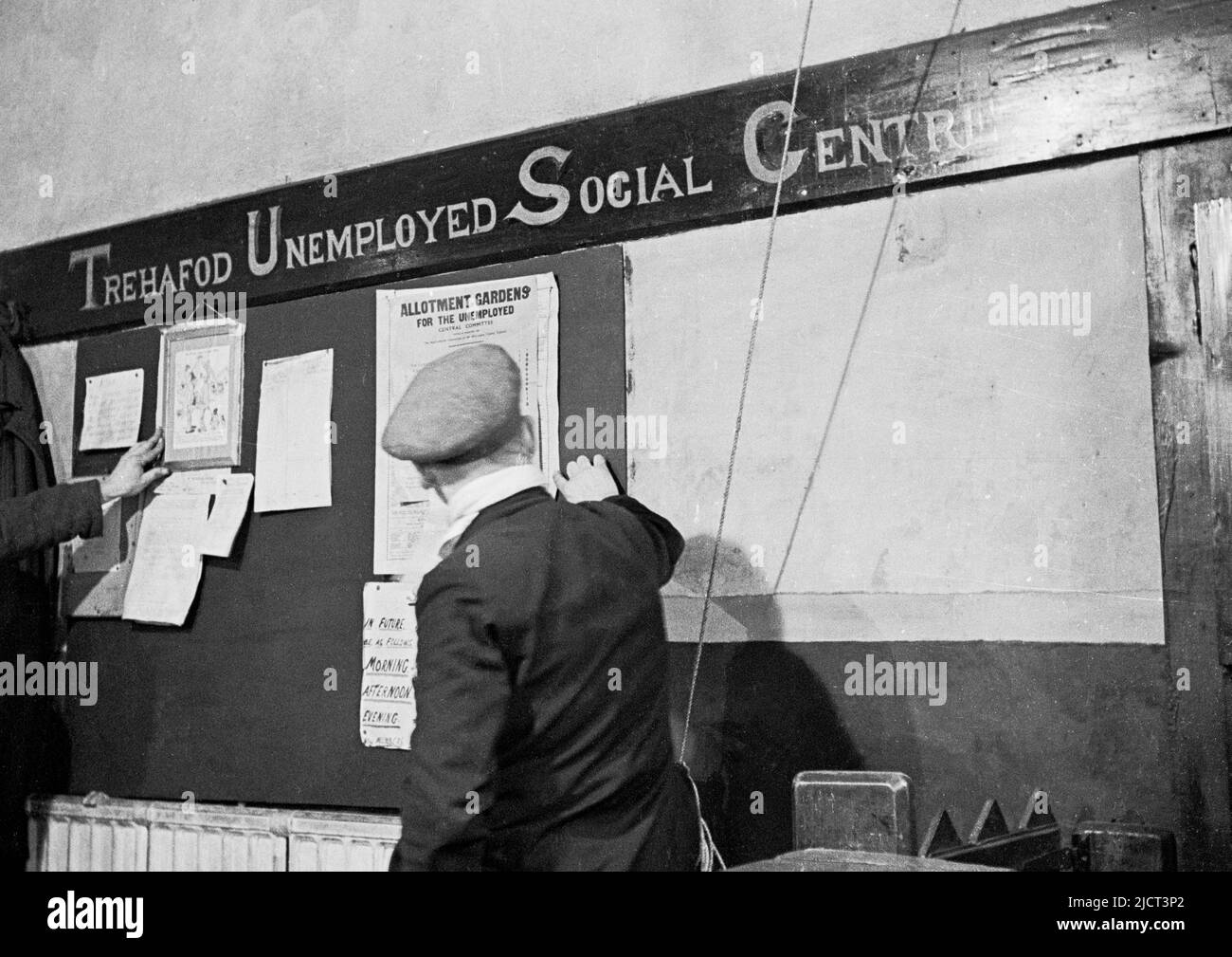 1940s, historique, un homme en tissu Cap regardant le tableau d'affichage au centre social de chômeurs de Trehafod. Trehafod, comme de nombreux villages du sud du pays de Galles, était tributaire de l'extraction du charbon, avec un manque d'opportunités de travail alternatives. Il y avait un chômage élevé à cette époque et un centre a donc été mis en place pour les mineurs sans emploi pour se rencontrer pour une discussion et un jeu de cribbage. L'un des avis lit, "les jardins d'allotement pour les chômeurs". Banque D'Images