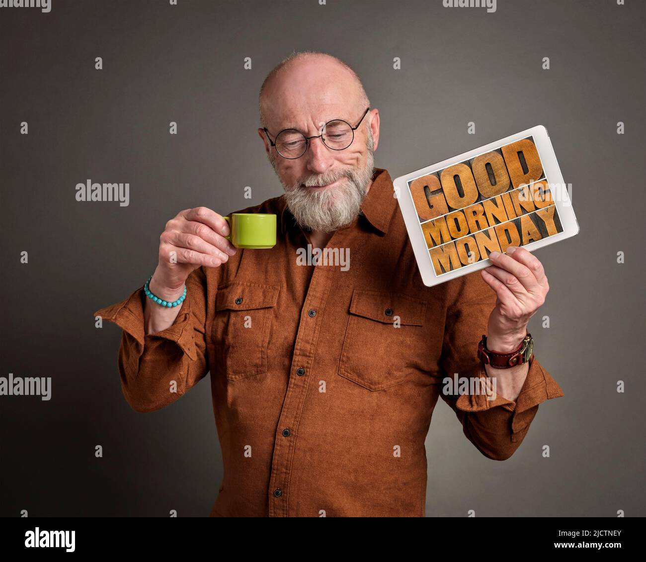 Bonjour lundi - un homme senior souriant boit du café et montre une tablette numérique avec du texte en bois, début positif d'une nouvelle semaine de travail co Banque D'Images