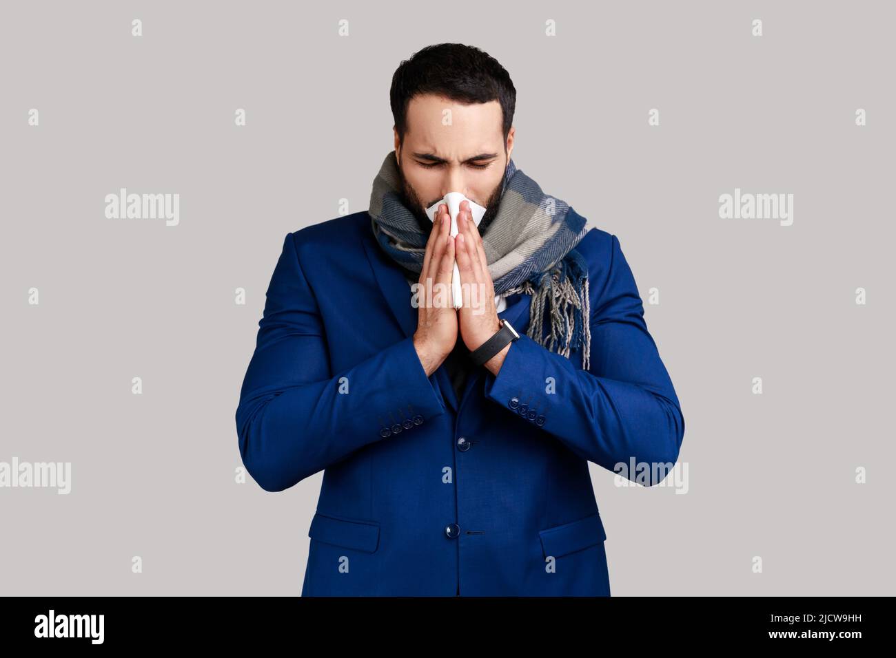 Homme enveloppé dans un foulard chaud éternuant dans la serviette, nettoyant le nez qui coule, souffrant de symptômes d'allergie ou de grippe, épidémie de grippe saisonnière, infection virale. Prise de vue en studio isolée sur fond gris. Banque D'Images