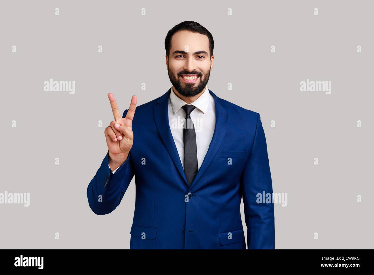 Homme barbu montrant le symbole du signe v de la paix avec les doigts, regardant la caméra avec le sourire crasseux, célébrant sa victoire réussie, portant le costume. Prise de vue en studio isolée sur fond gris. Banque D'Images