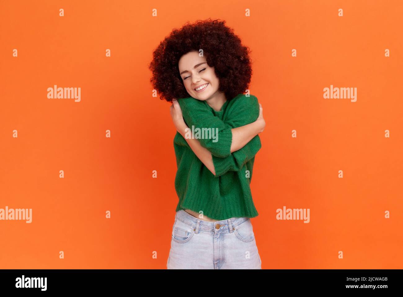 Femme à la coiffure afro portant un pull vert décontracté se embrassant et souriant, se sentant à l'aise et accomplie, narcissique personne égoïste. Studio d'intérieur isolé sur fond orange. Banque D'Images