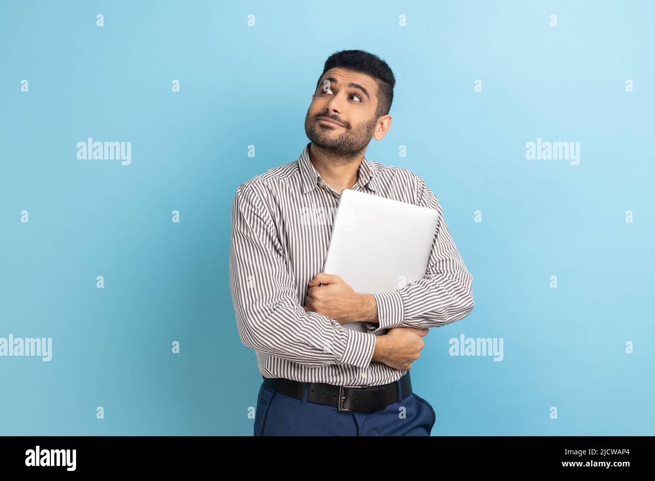 Portrait d'homme d'affaires pensif debout tenant un ordinateur portable fermé ou une chemise, regardant loin avec l'expression réfléchie, portant une chemise à rayures. Studio d'intérieur isolé sur fond bleu. Banque D'Images