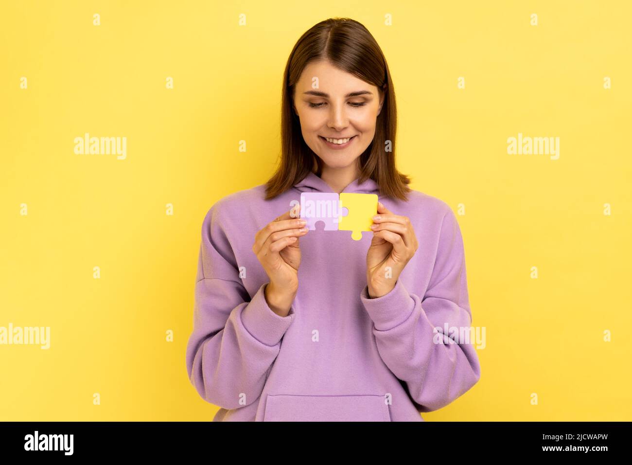 Portrait d'une femme attrayante et souriante, concentrée, tenant des pièces de puzzle jaunes et violettes, en train de résoudre des tâches, portant un sweat à capuche. Studio d'intérieur isolé sur fond jaune. Banque D'Images