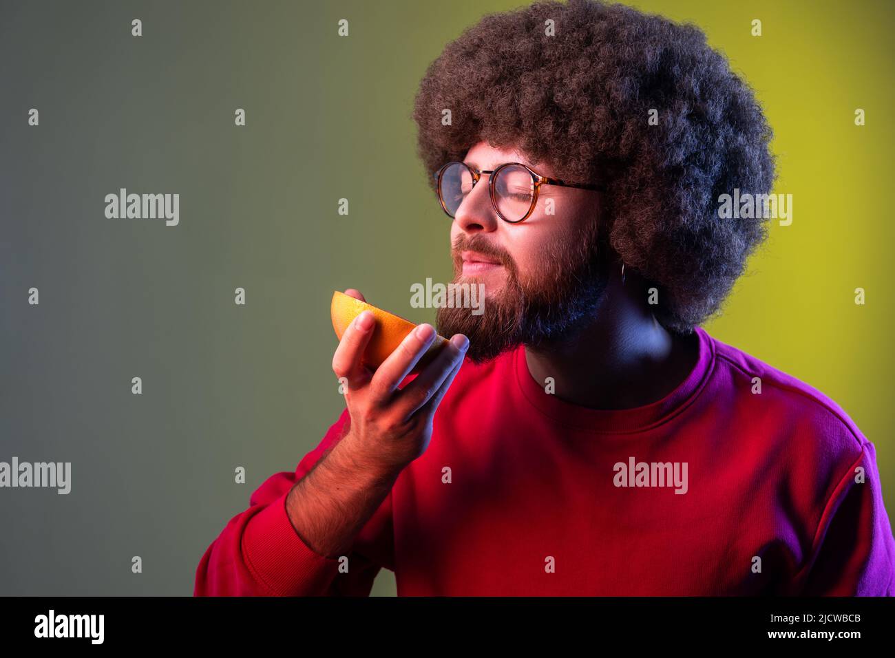 Hipster homme avec la coiffure d'Afro odorant demi tranche de pamplemousse, appréciant savoureux fruits frais juteux de vitamine, une alimentation saine. Studio d'intérieur isolé sur fond de néon coloré. Banque D'Images
