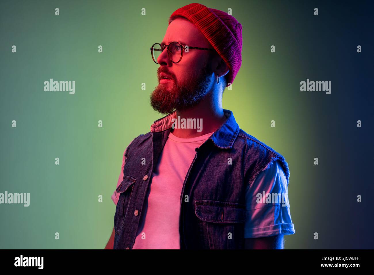 Vue latérale d'un joli boxeur avec barbe, beauté, portrait de mode, bonnet beanie et gilet en denim. Studio d'intérieur isolé sur fond de néon coloré. Banque D'Images
