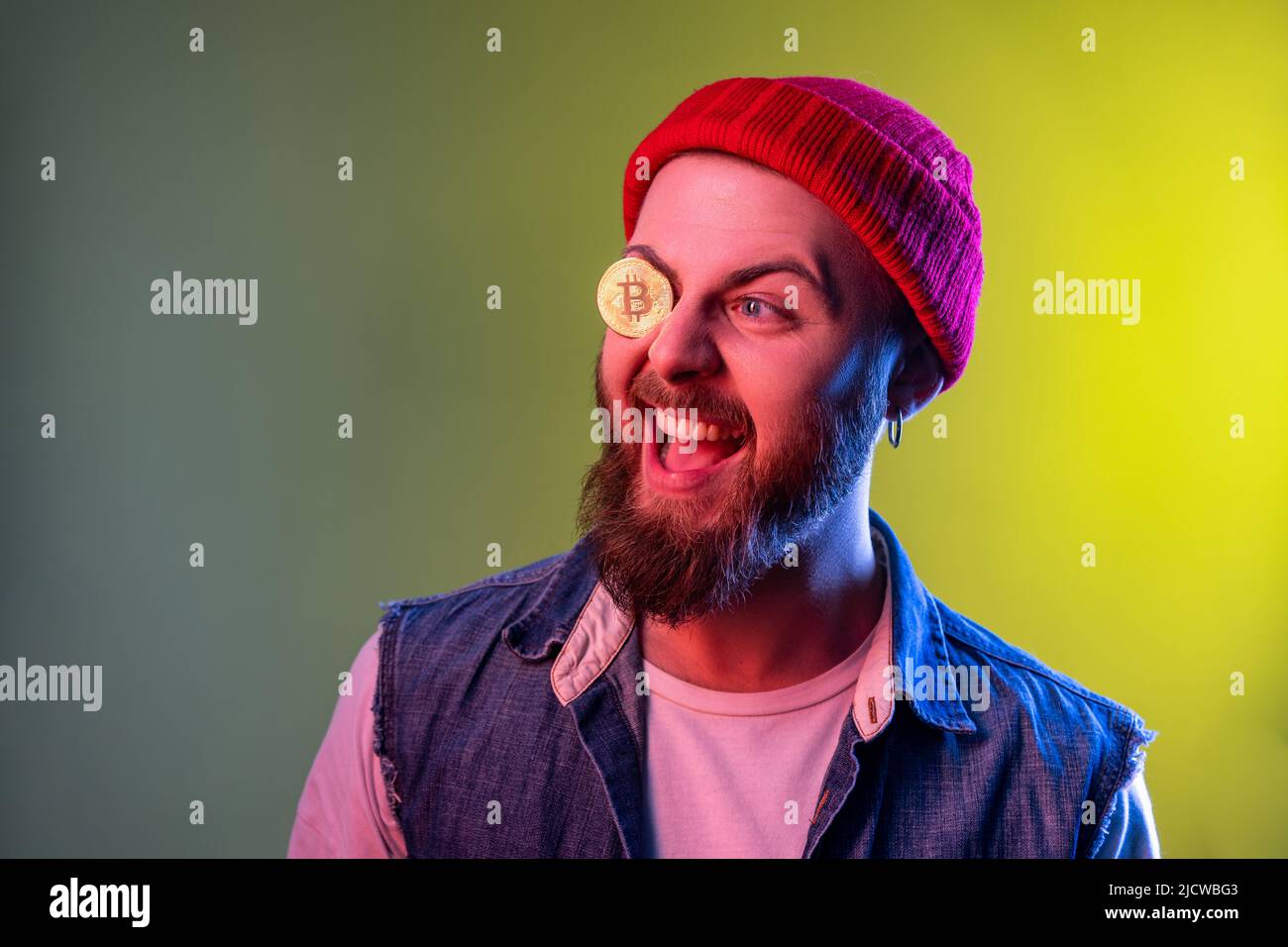 Joueur hippster homme debout couvrant un oeil avec le bitcoin doré, crypto-monnaie, regardant de côté, portant bonnet beanie et gilet en denim. Studio d'intérieur isolé sur fond de néon coloré. Banque D'Images