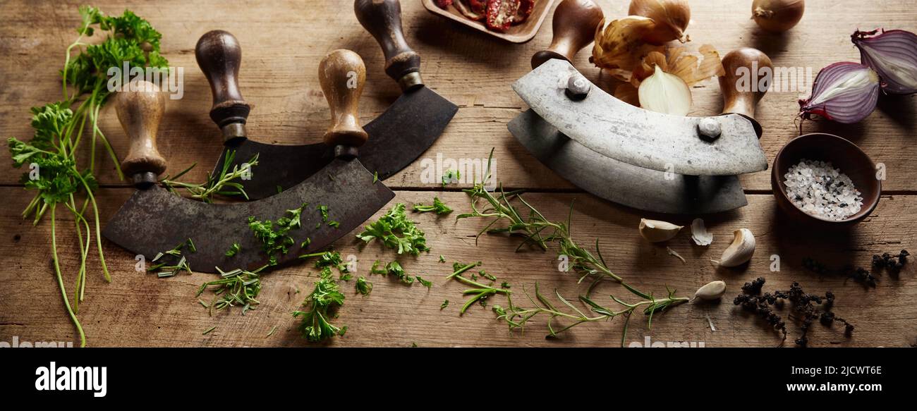 Depuis le dessus de la mezzaluna traditionnelle couteaux pointus placés sur une table en bois avec des herbes éparpillées pendant le processus de cuisson dans la cuisine légère Banque D'Images