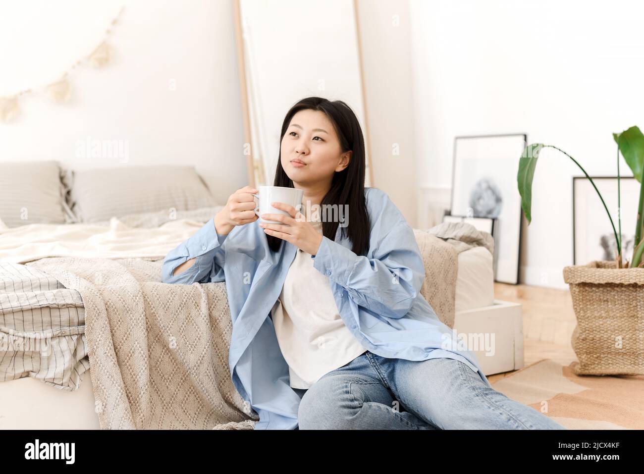 Photos d'une charmante jeune femme asiatique assise sur le sol près du lit, buvant du café et appréciant le week-end le matin. Banque D'Images