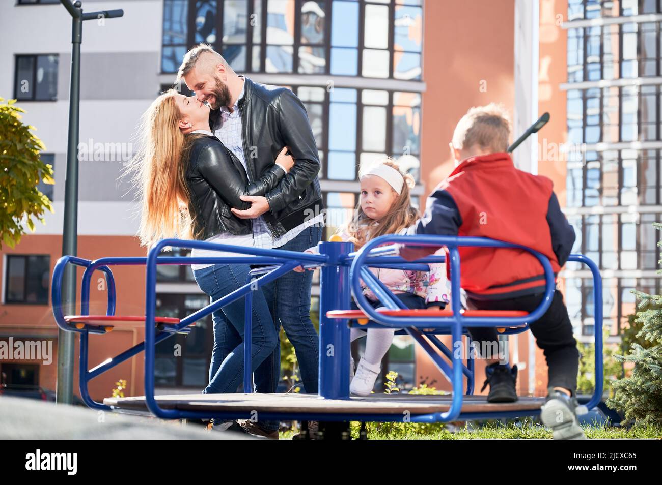 Les parents heureux traînaient avec les enfants, tandis que les enfants balançaient dans l'aire de jeux. Vue de face de deux adorables enfants qui tournaient autour du joyeux, tandis que les parents s'embrassant derrière l'extérieur. Concept de famille. Banque D'Images