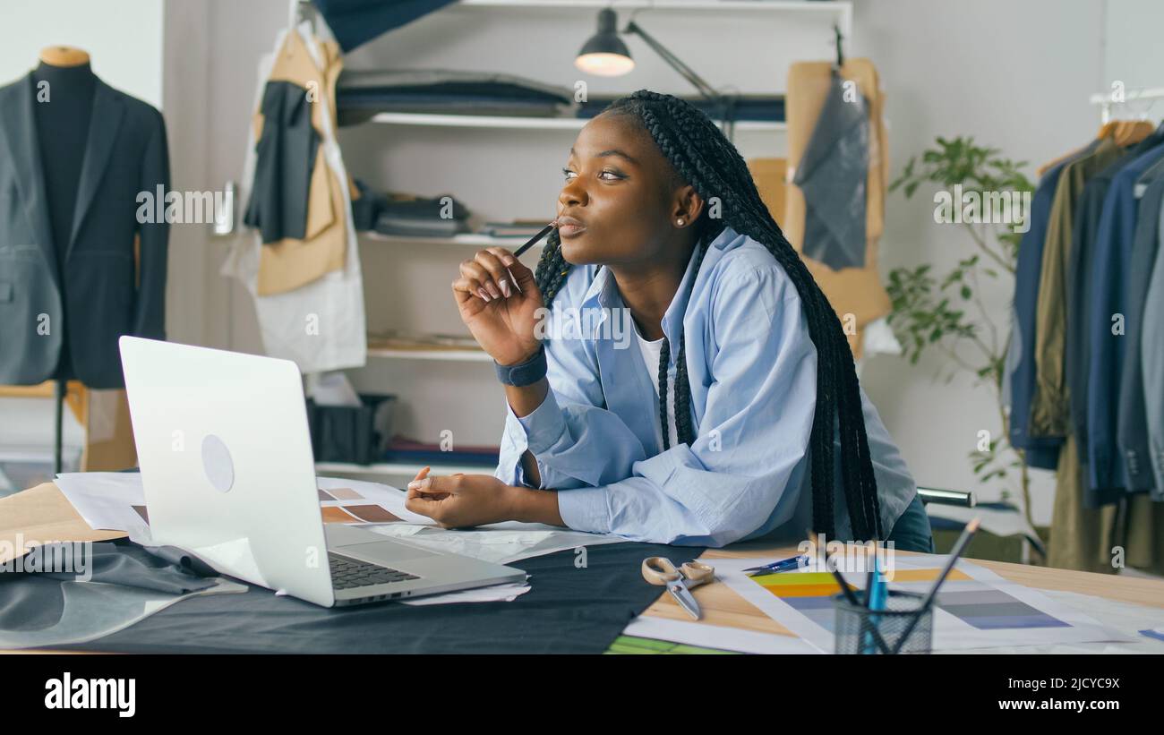 Un talentueux designer de mode afro-américain crée une nouvelle collection de mode tout en travaillant sur un ordinateur portable dans un studio Fashion Business Startup. Concept Banque D'Images